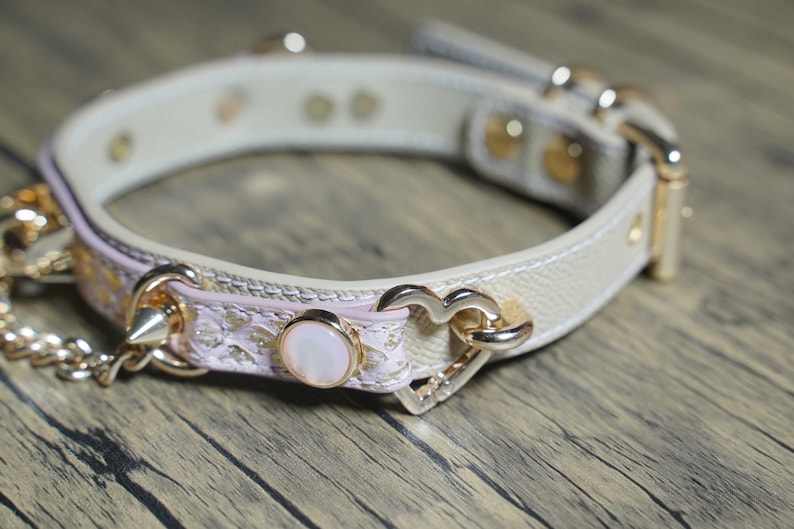 Puede incluir: Un collar gargantilla de cuero blanco con ribete de encaje rosa y detalles dorados. El collar presenta un dije en forma de coraz&oacute;n, un bot&oacute;n perlado y un detalle de punta.
