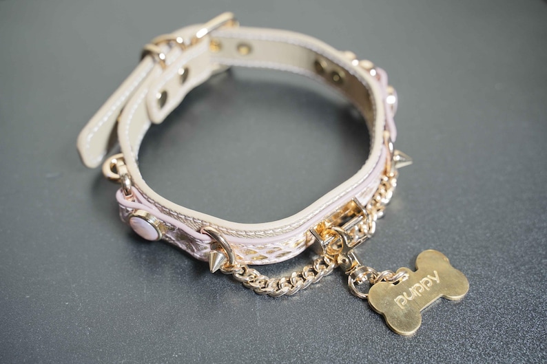 Puede incluir: Un collar de perro rosa y dorado con una cadena y una etiqueta en forma de hueso que dice "Puppy".