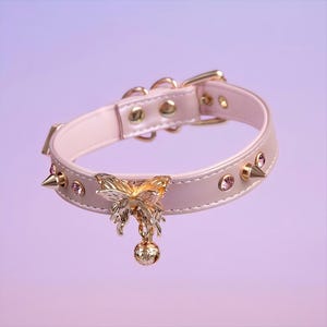May include: A pink leather choker with gold accents, featuring a butterfly charm and a bell. The choker is adorned with small pink rhinestones and gold spikes.