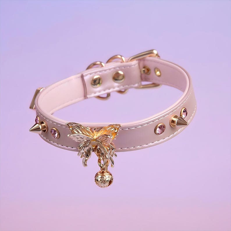 May include: A pink leather choker with gold accents, featuring a butterfly charm and a bell. The choker is adorned with small pink rhinestones and gold spikes.