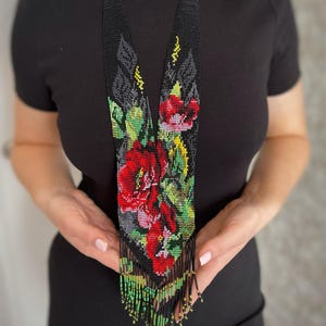 May include: A beaded necklace with a floral design. The necklace features a black background with red roses, green leaves, and yellow accents. The necklace has a fringe at the bottom and is held by a person wearing a black dress.