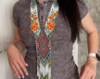 May include: A long beaded necklace featuring a V-shaped design with intricate geometric patterns in red, white, green, and yellow. The piece includes floral motifs at the top and a long fringe of white, green, and yellow beads at the bottom.