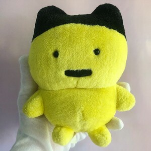 May include: A yellow plush toy with black ears and a black face. The toy has a simple, friendly expression with two black eyes and a small black mouth.