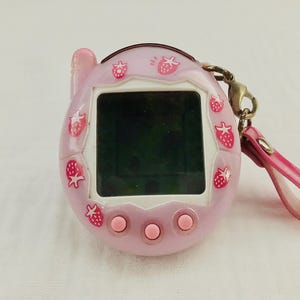 May include: A pink handheld electronic game with a black screen, adorned with strawberry graphics. It features three pink buttons and a pink strap. The device has a small antenna-like extension on top.