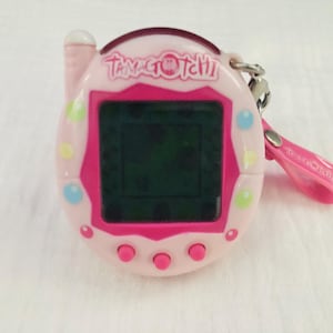 May include: A pink Tamagotchi digital pet with a black and white screen. The device has a pink strap and the text "Tamagotchi" is printed on the front.