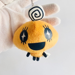 May include: A yellow plush toy with a black smiling face and sparkling eyes. The toy has a white swirl on top of its head and is attached to a keychain.