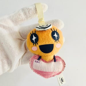 May include: A yellow plush toy with a black and white swirl on its head, black eyes with white stars, a black smiling mouth, and pink cheeks. The toy is holding a pink heart-shaped tag with the Japanese word "なまえ" written on it.