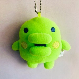 May include: A lime green plush toy with a black mouth and yellow eyes. The toy is shaped like a frog and has a chain attached to the top of its head.