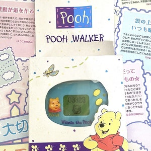 May include: A blue and white Winnie the Pooh character toy with a digital screen. The toy is in a box with a yellow background and a purple border. The box has a picture of Winnie the Pooh on it, as well as the text "Pooh Walker" and "Winnie the Pooh".