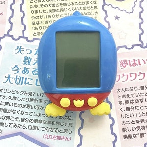 May include: A blue and red handheld electronic toy with a screen and buttons. The toy has a yellow beak and feet. The text on the toy reads "夢はいつだって ワクワクするも".