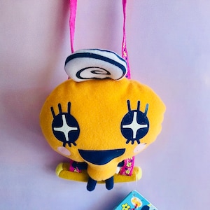 May include: A yellow plush toy with a white and black swirl on its head. The toy has large black eyes with white stars, a black smile, and pink cheeks. The toy is hanging from a pink strap.