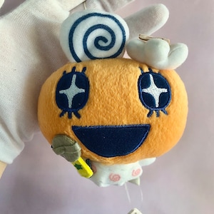 May include: A plush toy with a yellow body, blue eyes with white stars, and a blue smiling mouth. The toy has a white swirl on top of its head and a white tail with pink polka dots.