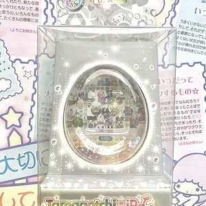 Könnte beinhalten: Ein weißes und silbernes Tamagotchi iD L Spielzeug in seiner Verpackung. Das Spielzeug ist eiförmig und hat einen bunten Bildschirm mit verschiedenen Figuren. Die Verpackung ist mit Sternen und Herzen verziert.
