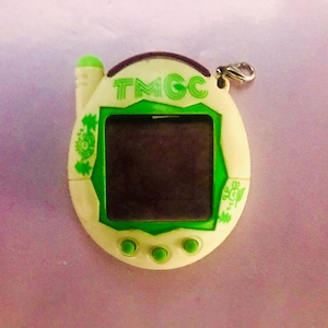 May include: A white and green digital pet toy with a screen and buttons. The toy has a green antenna and a silver clip. The text "TMGC" is printed on the toy.