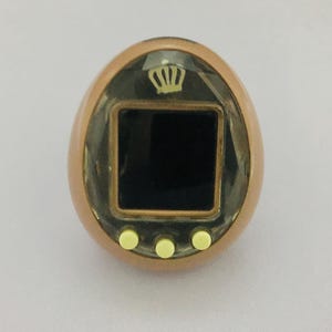 May include: A brown egg-shaped digital pet with a black screen and three white buttons. The screen has a gold crown icon.