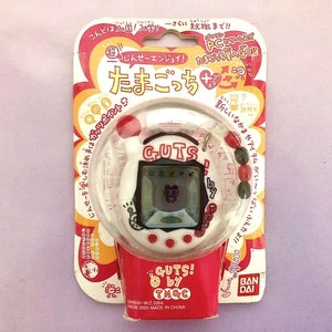 May include: White and red plastic Tamagotchi-like handheld electronic pet toy with a black and white screen. The toy is in its original packaging and features the text "GUTS! by TMGC" and "BANDAI".