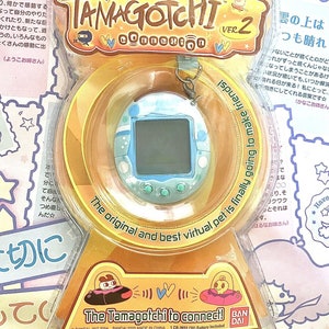 May include: A blue and green Tamagotchi Connection Ver. 2 virtual pet toy in a clear plastic package with a yellow background. The packaging features the text "The original and best virtual pet is finally going to make friends!" and "The Tamagotchi to connect!"