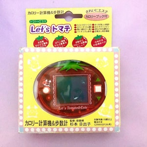 May include: A red and green plastic toy shaped like a tomato with a digital screen. The toy is labeled "Let's Tomato & Este" and has a calorie counter and pedometer. The box is yellow with white polka dots and says "Calorie Counter & Pedometer".