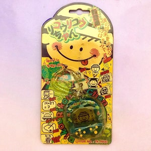 May include: A yellow and green plastic toy with a digital display and a chain. The toy is packaged in a yellow and green cardboard box with Japanese text. The text on the box reads "リフラリコンちゃん" and "ITEM NO. 8 TK: 9900".