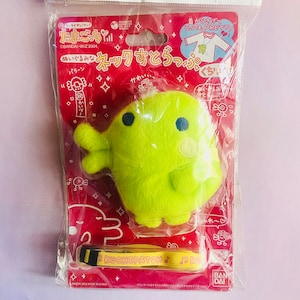 May include: A green plush toy with a yellow strap that says "KUCHIPATCH". The toy is packaged in a plastic bag with a red background and Japanese text. The text says "Tamagotchi" and "Earl Pretty".