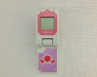 May include: A white and pink toy phone with a flip-up screen. The screen has a pink border and says "Pretty Cure" on it. The phone has a purple bottom with a pink rose design.