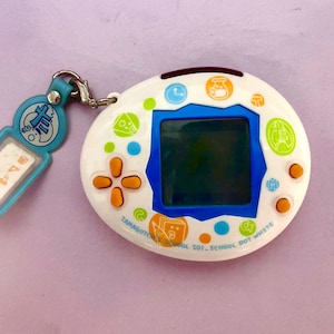 May include: A white Tamagotchi with a blue screen and orange buttons. The device has a white and blue strap with a small blue tag that says "T.M & C. SCHOOL". The Tamagotchi is decorated with colorful dots and icons, including a clock, a beaker, and a book.