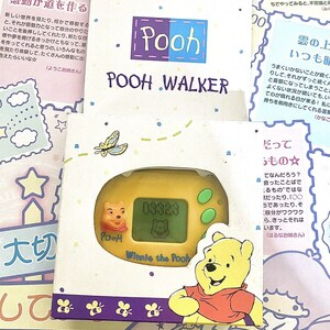 May include: A yellow Winnie the Pooh character handheld electronic game with a digital display. The game is in a box with a purple, yellow, and white design. The box has the text "Pooh Walker" on it.