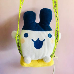 Könnte beinhalten: Ein Plüschtier mit weißem Körper, blauen Ohren und einem blauen lächelnden Gesicht. Das Spielzeug ist an einem grün-weißen Band mit rosa Blumen befestigt. Das Spielzeug sitzt auf einem gelben Kissen.