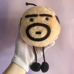 May include: A soft toy with a circular, beige body and black facial features. It features eyebrows, eyes, and a mouth. It has a small, brown topknot and two dangling legs with black pom-pom feet. The toy is held by a person wearing a white glove.
