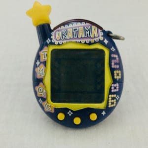 May include: A blue and yellow Tamagotchi with a star on top. The screen is black and the text on the front reads "URATAMA".