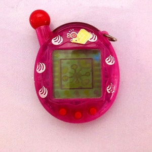 Puede incluir: Un juego de mascotas digital rosa con un botón rojo y una bola roja en la parte superior. La pantalla muestra un gráfico verde y blanco. El dispositivo tiene un pequeño anillo plateado adjunto en la parte superior. Diseños de remolino blanco decoran los lados.