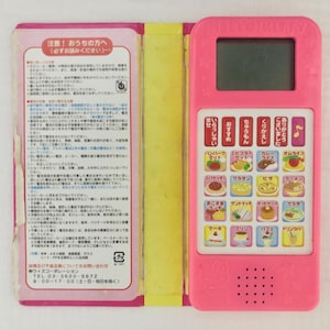 Pode incluir: Um livro de brinquedo rosa e amarelo para crianças com um dispositivo eletrônico embutido. O dispositivo tem uma pequena tela e botões com imagens de diferentes alimentos. O texto no livro diz "注意! おうちの方へ (必ずお読みください)" e "ウィズコーポレーション TEL.03-3620-5672 8:00-17:00(土・日)".