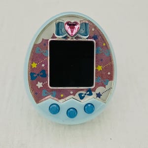 Puede incluir: Un juego electrónico portátil azul claro con una gema rosa en forma de corazón y una pantalla negra. El dispositivo tiene tres botones azules en la parte inferior y un patrón decorativo de lazos, estrellas y nubes.