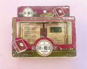 May include: A red and gold box with a picture of a book on the front. The box is labeled "The Oraclebook: Love Magic Wand Mobile" and features Japanese text. The box is for a digital fortune telling device.