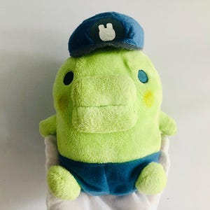 Puede incluir: Un juguete de peluche verde con un sombrero azul y un conejito blanco en el sombrero. El juguete lleva pantalones azules y tiene una gran sonrisa.