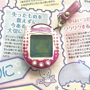 May include: A pink and white Tamagotchi digital pet toy with a white screen and pink buttons. The toy is on a pink and white background with Japanese text that reads "Lost things are not counted, but what is present is precious."