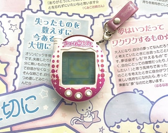 May include: A pink and white Tamagotchi digital pet toy with a white screen and pink buttons. The toy is on a pink and white background with Japanese text that reads "Lost things are not counted, but what is present is precious."