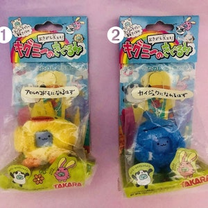 Puede incluir: Dos paquetes de juguetes de peluche con un diseño de animal de dibujos animados. Los paquetes están etiquetados con el texto "キノミーのそこあ" y "おきがえ犬スキ!" Los juguetes son amarillos y azules y tienen un aspecto lindo y de dibujos animados.