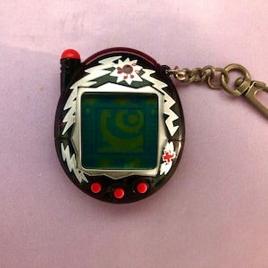 May include: Black and silver digital pet with a red antenna and a silver chain. The screen shows a green and blue image. The pet has four red buttons.