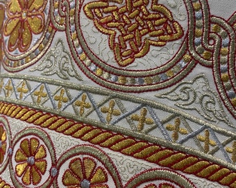 May include: Close-up of a white fabric with gold and red embroidered details. The design features a repeating pattern of circles with floral motifs and a rope-like border.