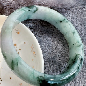 May include: A green jade bangle bracelet with a white background. The bracelet has a light green colour with darker green swirls throughout.