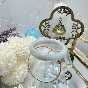 May include: A light green jade bangle bracelet resting on a clear glass surface with a gold rim. The bracelet is surrounded by white flowers and a decorative gold metal stand with a bell hanging from it.