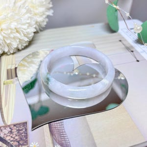 May include: A white jade bangle bracelet with a light green hue. The bangle is displayed on a heart-shaped mirror.