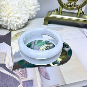 May include: A light blue jade bangle bracelet displayed on a heart-shaped mirror with a floral design. The bangle is smooth and polished, with a subtle sheen. The mirror is surrounded by a white frame.