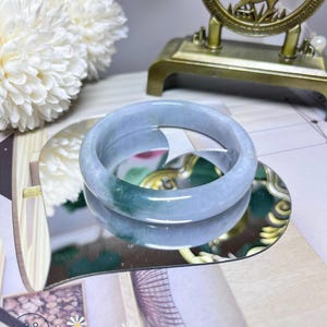 May include: A light blue jade bangle with a green hue. The bangle is displayed on a heart-shaped mirror with a floral design.