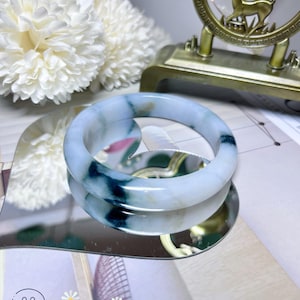 May include: A jade bangle bracelet in shades of light blue and green. The bracelet is reflected in a mirror. The image also includes white flowers, a gold-coloured decorative object, and the text "Chicago Jade".