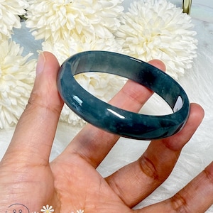 May include: A polished, dark teal jade bangle bracelet. The smooth, rounded bracelet is held in a hand, showcasing its glossy surface. The logo "Chicago Jade" is visible in the lower left corner.