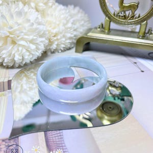 May include: A light blue jade bangle bracelet on a heart-shaped mirror. The bracelet is smooth and polished. The mirror is sitting on a book with the text "Chicago Jade" printed on it.