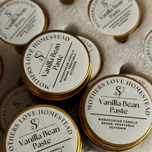 May include: Close-up of several small, round containers of vanilla bean paste. Each container has a white label with gold trim, featuring the text "Mothers Love Homestead" and "Vanilla Bean Paste." The ingredients are also listed.