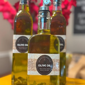 May include: Three clear glass bottles filled with olive oil, each with a metal spout and a label that reads "Olive Oil 100% Extra Virgin Garlic Infused." The bottles are on a yellow surface, with red flowers in the background.